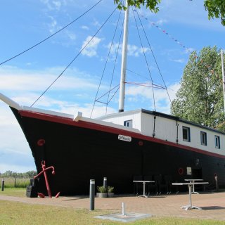 Das historische Hotelschiff „Stinne“, ein ehemaliger Küstenschoner
                           mit schwarzem Rumpf und weißem Decksaufbau, steht fest an Land im Ostseebad Wustrow. Am Bug hängt ein markanter, rot gestrichener
                           Anker, und am hohen Mast ist eine bunte Wimpelkette befestigt. Im Vordergrund lädt eine gepflasterte Terrasse mit weißen Tischen
                           zum Verweilen unter blauem Sommerhimmel ein.