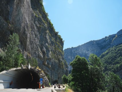 Mehrere Personen mit Fahrrädern fahren
                           bzw. stehen auf einer Straße vor einem Tunnel in einer hohen Felslandschaft. Links befindet sich der Tunneleingang, darüber
                           und daneben ragen steile Kalkfelsen auf. Im Vordergrund sind Straße, Verkehrsschilder und Bäume zu sehen.