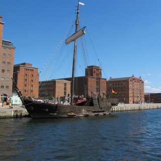Ein historisches
                           Holzsegelschiff, eine Hansekogge, liegt im Alten Hafen von Wismar vor Anker. Im Hintergrund ragen große, restaurierte Speicherbauten
                           aus rotem Backstein unter einem wolkenlosen, blauen Himmel empor. Das ruhige, blaue Hafenwasser spiegelt die Szenerie wider.