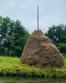Ein großer kegelförmiger Heuhaufen
                           steht auf einer grünen Wiese am Ufer eines schmalen Wasserlaufs. Aus der Spitze ragt ein langer Holzpfahl. Im Hintergrund
                           wachsen dichte Laubbäume unter einem bedeckten Himmel.