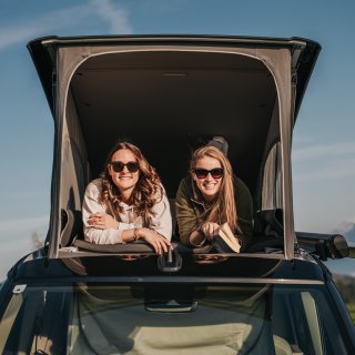 Zwei Personen liegen im geöffneten Aufstelldach eines Campers und blicken nach vorne. Eine Person hält ein Buch in der
                           Hand. Das Fahrzeug steht im Freien unter blauem Himmel.