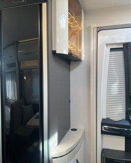 Innenansicht des Hobby
                           Excellent 460 SL mit hellem Schrankelement, spiegelnder dunkler Fläche und einem Fenster mit geschlossenem Rollo. Rechts unten
                           ist ein Teil eines Möbelblocks zu sehen, links spiegelt sich der Wohnbereich mit Sitzgruppe und Schlafbereich.