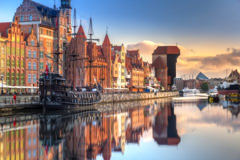 Die Danziger Altstadt in
                           der Abendsonne, die bunten Häuserfassaden spiegeln sich im Wasser der Mottlau, ein altes Segelschiff in der linken Bildhälfte
                           sowie das bekannteste Wahrzeichen der Stadt, das Krantor, sind zu sehen.