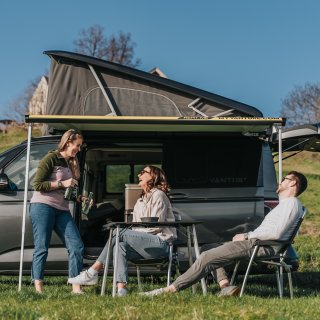 Drei Personen sitzen und stehen vor einem Camper mit ausgefahrener Markise und geöffnetem Aufstelldach.
                           Vor dem Fahrzeug befinden sich ein Campingtisch und Stühle auf einer Wiese.