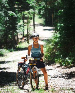 Eine Person mit Fahrradhelm steht
                           mit einem bepackten Fahrrad auf einem steinigen Waldweg. Der Weg verläuft zwischen dichtem Grün und Bäumen, im Hintergrund
                           ist ein hellerer Abschnitt des Weges zu sehen.