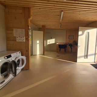 Holzverkleideter Vorraum
                           des Sanitärgebäudes mit zwei Waschmaschinen, mehreren Türen und einer Wickelkommode im hinteren Bereich.