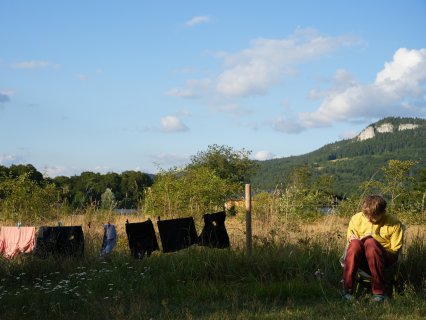 Eine Person sitzt auf einem Campingstuhl
                           in einer Wiese und schreibt in ein Heft. Daneben hängt Wäsche auf einer Leine. Im Hintergrund sind Sträucher, Bäume, bewaldete
                           Hügel und helle Felswände unter blauem Himmel zu sehen.