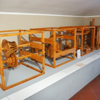 Mehrere
                           mechanische Holzmodelle sind in einer Reihe auf einem langen, hellgrauen Podest ausgestellt. Die Konstruktionen bestehen aus
                           massiven, hellen Holzrahmen, in denen unterschiedliche technische Elemente wie Zahnräder, Antriebswellen, Speichen und Hebel
                           verbaut sind. Auf dem Podest vor den Apparaturen liegen flache, weiße Informationstafeln mit Texten und historischen Skizzen.
                           Der Raum ist schlicht gehalten, mit einer hellbeigen Wand im Hintergrund und quadratischen, rötlich-braunen Fliesen auf dem
                           Boden.