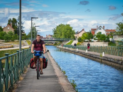 Eine Person fährt mit einem bepackten Fahrrad auf
                           einem befestigten Weg entlang eines Kanals. Auf der gegenüberliegenden Seite sind weitere Personen mit Fahrrädern, Geländer,
                           eine Brücke, Bäume und Häuser zu sehen. Der Himmel ist blau mit mehreren Wolken.