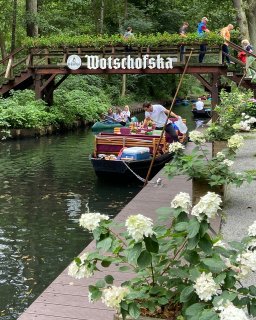 Ein Spreewaldkahn mit roten Polstern liegt an einem Holzsteg vor der Wotschofska-Brücke.
                           Eine Person bereitet den Kahn am Ufer vor, während weitere Personen die mit Blumen geschmückte Holzbrücke überqueren. Hinter
                           dem Kahn fahren mehrere Personen in Kanus auf dem schmalen Wasserlauf.