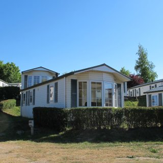 Moderne,
                           helle Mobilheime im Ostseecamp Seeblick in Rerik. Die Ferienhäuser stehen leicht erhöht auf einer Anhöhe und sind mit Erkern,
                           großen Fenstern und dunklen Fensterläden ausgestattet. Eine gepflegte, grüne Hecke trennt die Unterkünfte vom davorliegenden
                           Weg unter blauem Himmel.