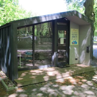 Ein
                           moderner, anthrazitfarbener Büro-Container mit einer großen Glasfront und einer kleinen Holzterrasse im schattigen Wald des
                           Campingplatzes. Neben der verglasten Eingangstür hängt ein gelbes Schild mit der Aufschrift „Ihr mietbarer OSTSEE ARBEITS
                           PLATZ“. In den Fensterscheiben spiegeln sich die umliegenden Bäume.