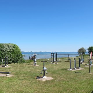 Ein moderner Outdoor-Fitnessparcours
                           auf einer grünen Rasenfläche im Ostsee-Campingpark Zierow. Verschiedene graue und schwarze Trainingsgeräte stehen verteilt
                           auf der Wiese, die durch einen Zaun vom dahinterliegenden, ruhigen Meer getrennt ist. Über der Szenerie wölbt sich ein strahlend
                           blauer Himmel.