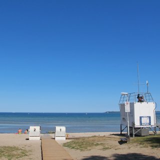 Der flach abfallende Sandstrand
                           am Ostsee-Campingpark Zierow unter strahlend blauem Himmel. Ein Holzsteg führt im Vordergrund in Richtung Wasser, vorbei an
                           zwei weißen Strandkörben mit den Nummern 1 und 2. Auf der rechten Seite steht eine weiße Rettungsschwimmer-Station der DLRG.