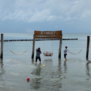 Eine hölzerne Schaukel- und Hängemattenkonstruktion
                           direkt im flachen Ostseewasser am Strand von Zingst. Am oberen Holzbalken stehen die Schriftzüge „ZINGST“ und „HALB INSEL,
                           HALB PARADIES“. Im Wasser stehen zwei Erwachsene, während zwei kleine Kinder in der geflochtenen Schaukel sitzen. Der Himmel
                           im Hintergrund ist dicht bewölkt.