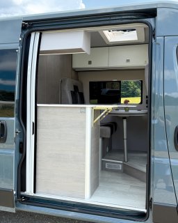 Blick von außen durch die geöffnete Schiebetür in den Innenraum des Campers. Deutlich zu sehen ist die seitliche
                           Verlängerung der Küchenzeile, die in den Türbereich ragt, sowie der Durchblick zur Sitzgruppe.