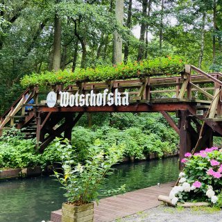 Dunkelbraune Holzbrücke mit der weißen
                           Aufschrift „Wotschofska“ über einem schmalen Wasserlauf. Blumenkästen mit roten Blüten schmücken das Brückengeländer; am Ufer
                           stehen Pflanzgefäße mit weißen und rosafarbenen Hortensien. Dichte grüne Bäume und Sträucher umgeben die Brücke.