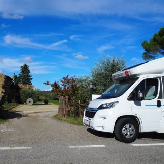 Ein weißes Wohnmobil steht
                           auf der rechten Seite an einer asphaltierten Straße im hellen Sonnenschein. Links daneben öffnet sich eine geschotterte Einfahrt,
                           die an einem einfachen Holzzaun und mehreren Olivenbäumen vorbei zu einem rustikalen Steinhaus mit Ziegeldach führt. Im weitläufigen
                           Gartenbereich davor liegt ein großer, aufgestellter Mühlstein. Die ländliche Szenerie liegt unter einem strahlend blauen Himmel
                           mit weißen Schönwetterwolken.