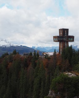 Jakobskreuz auf der bewaldeten Buchsteinwand vor herbstlicher Landschaft
                           und verschneiten Bergen.
