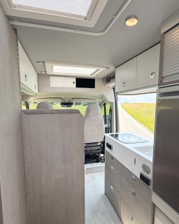 Blick vom hinteren Bereich in Richtung Fahrerhaus. Rechts befindet sich die Küchenzeile mit Herd und Spüle, darunter
                           sind mehrere Schubladen in Holzoptik verbaut. Links ist ein hohes Möbelstück in Holzdekor zu sehen, im Hintergrund die Sitze
                           des Fahrerhauses.