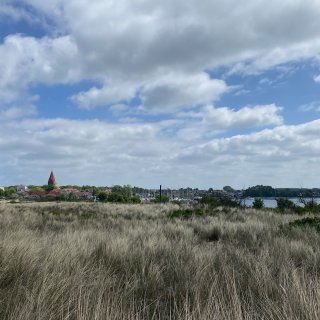 Blick über eine
                           weite, von trockenem Dünengras geprägte Landschaft auf das Ostseebad Rerik. Im Hintergrund ist die Silhouette des Ortes mit
                           dem markanten roten Turm der St.-Johannes-Kirche und dem angrenzenden Salzhaff zu sehen, an dessen Ufer Segelboote liegen.
                           Über der Szenerie erstreckt sich ein blau-weißer Wolkenhimmel.