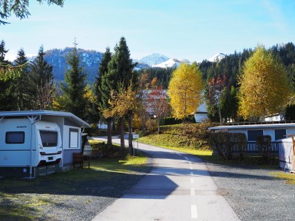 Im Bild sieht man den Eingang zum Tirol Camp Fieberbrunn. Es ist eine kleine Straße mit Bäumen links und
                           rechts, sowie einen Wohnwagen, welcher rechts steht. Im Hintergrund sieht man ein Bergpanorama.