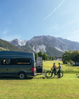 Zwei Personen stehen mit Fahrrädern neben einem Camper auf einer Wiese. Im Hintergrund sind Berge, Bäume
                           und einzelne Gebäude zu sehen.