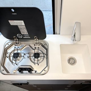 Draufsicht
                           auf den Küchenblock mit einem zweiflammigen Gaskocher und einem weißen Spülbecken. Der Kocher verfügt über eine hochgeklappte,
                           schwarze Abdeckung mit Sicherheitshinweisen.