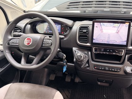 Blick auf das dunkelgraue
                           Armaturenbrett des Camper-Vans. Links befindet sich das Multifunktionslenkrad mit rotem Fiat-Logo, dahinter die teilweise
                           digitale Instrumententafel. In der Mittelkonsole sticht der Schaltknüppel für das manuelle 6-Gang-Getriebe hervor, der Schlüssel
                           steckt im Zündschloss. Dominant in der Mitte ist ein großes Touch-Display, das gerade das Bild der Rückfahrkamera aus dem
                           hinteren Fahrzeugbereich zeigt. Darunter sind die Bedienelemente der Klimaanlage und weitere Fahrzeugtasten angeordnet. Unten
                           links ist ein Teil des Fahrersitzes angeschnitten.