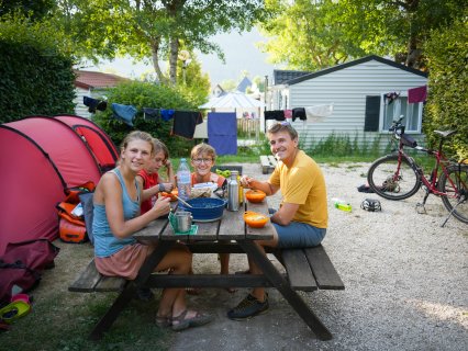 Vier Personen sitzen an einem Picknicktisch auf
                           einem Campingplatz und essen. Neben dem Tisch stehen ein Zelt und zwei Fahrräder, im Hintergrund sind weitere Unterkünfte
                           und eine Wäscheleine zu sehen.