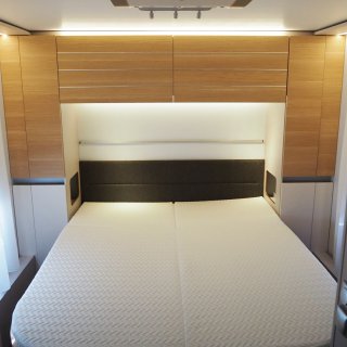 Beleuchteter Schlafbereich/Doppelbett
                           im Inneren des Wohnwagens Adria Adora 593 UP mit Holzkästen darüber und daneben.