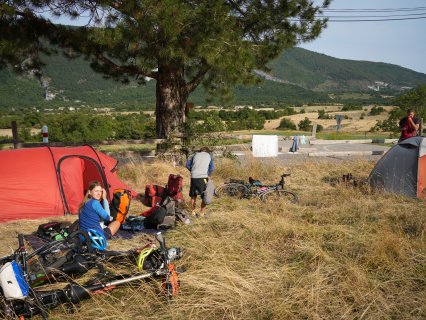 Mehrere Zelte stehen auf einer
                           trockenen Wiese unter einem großen Baum. Daneben liegen bepackte Fahrräder, Taschen und Campingausrüstung. Im Hintergrund
                           sind eine Straße, Wäsche auf einer Leine, Felder und bewaldete Berghänge zu sehen.