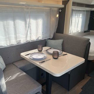 Sitzgruppe im Hobby Excellent 460 SL mit grauer Polsterbank, rechteckigem
                           Tisch und eingedecktem Geschirr für zwei Personen. Neben dem Tisch befinden sich ein großes Fenster mit hellen Vorhängen,
                           ein Wandlicht und im Hintergrund ein Teil des Schlafbereichs.
