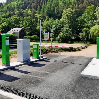 Einfahrt von CAMPING-CAR PARK mit Schranke, Wald und kleines Haus im Hintergrund.