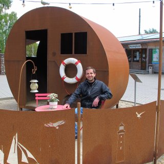 Ein lächelnder Mann in einer dunklen
                           Jacke sitzt an einem kleinen rosa Tisch vor einer außergewöhnlichen, runden Unterkunft aus rostbraunem Metall. An der kabinenartigen
                           Konstruktion hängt ein rot-weißer Rettungsring, davor schützt ein niedriger Metallschnitt-Zaun mit maritimen Motiven wie einem
                           Leuchtturm und Möwen den Sitzbereich. Im Hintergrund ist das Rezeptionsgebäude des Campingplatzes erkennbar.
