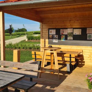 Überdachte Holzterrasse mit Sitzbänken,
                           Tischen, einem Stehtisch und einer schwarzen Informationstafel an der Wand.