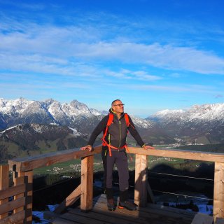 Person mit Rucksack auf einer hölzernen Aussichtsplattform vor einem teilweise
                           verschneiten Bergpanorama.