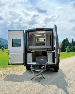 Ein grauer Camper-Van steht mit weit geöffneten
                           Hecktüren auf einer kleinen Landstraße vor einer saftig grünen Wiese. Man blickt direkt in den hellen Innenraum mit Möbeln
                           in Holzoptik und einem teilweise angehobenen Bett, während draußen vor dem Heck ein grauer Campingstuhl für eine entspannte
                           Pause bereitsteht.