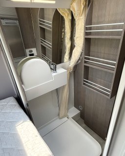 Blick auf den Waschraum, bei dem das Waschbecken nach oben geklappt wurde, um die Duschwanne freizugeben. Rechts
                           sind die offenen Regale in Holzoptik zu sehen, links grenzt die Wandverkleidung an den Schlafbereich an.
