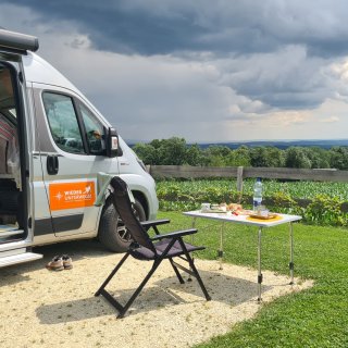 Campingbus auf einer geschotterten
                           Stellfläche mit Campingsessel, gedecktem Tisch, Rasen und weitem Blick über Felder und bewaldete Hügel.