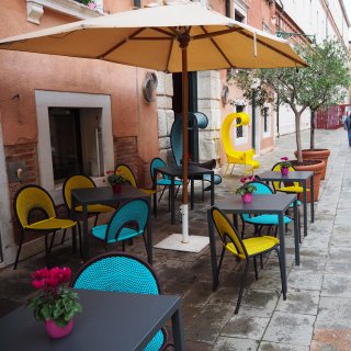 Ein einladender Außenbereich auf einer gepflasterten Gasse vor der terrakottafarbenen Fassade des
                           Ca' Pisani Hotels in Venedig. Unter einem großen, beigefarbenen Sonnenschirm gruppieren sich moderne Stühle in leuchtendem
                           Gelb und Türkis um dunkelgraue Tische, auf denen kleine pinke Alpenveilchen stehen. Im Hintergrund ziehen zwei extravagante,
                           hochgeschwungene Sessel die Blicke auf sich. Die Szenerie wird auf der rechten Seite durch junge Olivenbäume in großen Pflanzkübeln
                           abgerundet.