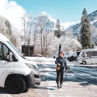 Frau mit Ski und Skistöcken in der Hand vor Reisemobil im Winter mit weiteren Reisemobilen auf Plätzen
                           sowie Bergpanorama im Hintergrund.