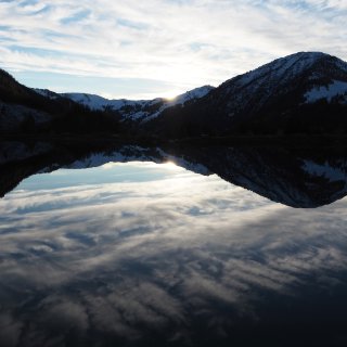 Ruhiger Bergsee, in dem sich verschneite Berghänge, die
                           tief stehende Sonne und Wolken spiegeln.