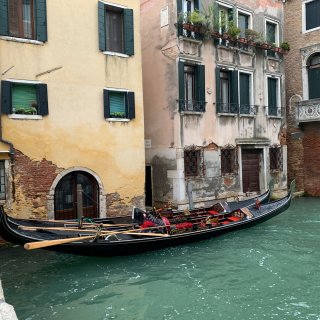 Zwei klassische, tiefschwarz lackierte
                           Gondeln mit kunstvollen goldenen Verzierungen und roten Polstersitzen ruhen nebeneinander vertäut in einem Kanal mit türkisgrünem
                           Wasser. Die Boote liegen vor historischen, verwitterten Hausfassaden, bei denen stellenweise der rote Backstein unter dem
                           abblätternden Putz hervortritt. Die Gebäude sind mit dunkelgrünen Fensterläden, schmiedeeisernen Gittern und kleinen, bepflanzten
                           Balkonen versehen.