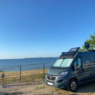 Ein grauer
                           Kastenwagen-Campingbus steht auf einem Stellplatz direkt an der Küste des Ostsee-Campingparks Zierow. Durch einen Metallzaun
                           getrennt, blickt man direkt auf den Sandstrand und das weite, blaue Meer der Wismarer Bucht unter einem wolkenlosen Himmel.
                           Am linken Bildrand ist eine Stromsäule zu sehen.