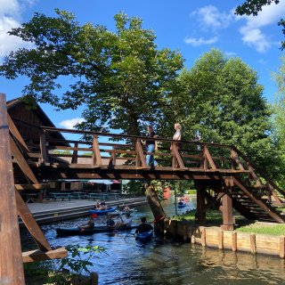 Zwei Personen gehen über
                           eine hohe Holzbrücke, während mehrere Personen mit Kajaks und Kanus auf dem schmalen Wasserlauf darunter hindurchfahren. Am
                           Ufer stehen Bäume und ein Gebäude mit Sitzplätzen im Freien.