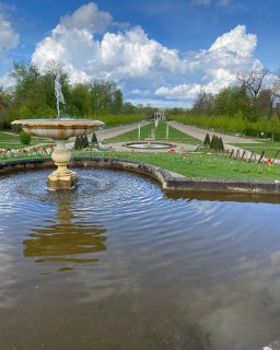 Ein historischer Springbrunnen
                           im weitläufigen, barocken Schlossgarten von Neustrelitz. Im Vordergrund plätschert Wasser in einem großen Rundbecken, dahinter
                           erstreckt sich eine symmetrisch angelegte Parkanlage mit grünen Rasenflächen, Tulpenbeeten und Alleen unter einem blau-weißen
                           Wolkenhimmel.