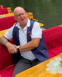 Eine Person mit
                           Brille, weißem Poloshirt und dunkelblauer Weste sitzt in einem Spreewaldkahn mit roten Polstern und Holztischen. Im Hintergrund
                           ist das grüne Wasser eines Fließes zu sehen.