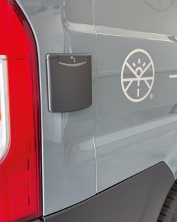 Nahaufnahme der Karosserie eines grauen Camper-Vans.
                           Direkt neben dem roten Rücklicht befindet sich eine kleine schwarze Klappe mit einem Wasserhahn-Symbol, hinter der sich der
                           Frischwasseranschluss verbirgt. Rechts daneben ist das runde weiße Herstellerlogo aufgedruckt.