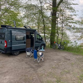 Ein grauer Kastenwagen-Campingbus steht auf
                           einer schattigen, von Bäumen umgebenen Stellplatz-Terrasse direkt am Seeufer. Davor sitzt eine Person auf einem Campingstuhl
                           an einem Tisch und liest ein Buch. Im Hintergrund lehnen Fahrräder an einem Baum am Hang mit Blick auf das Wasser.